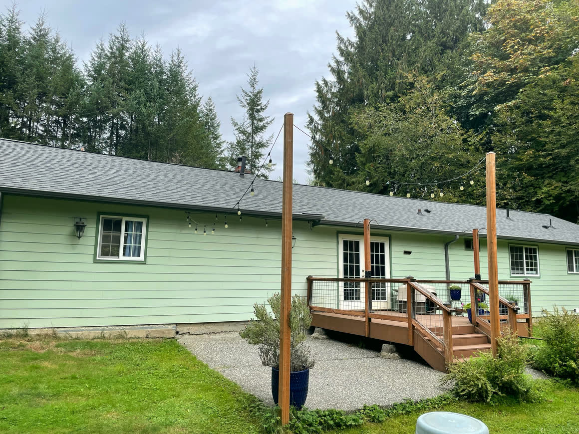 The rear of the house, sage-green lap siding under a grey shingle roof, with festoon lights strung across the wood deck and tall firs standing behind.