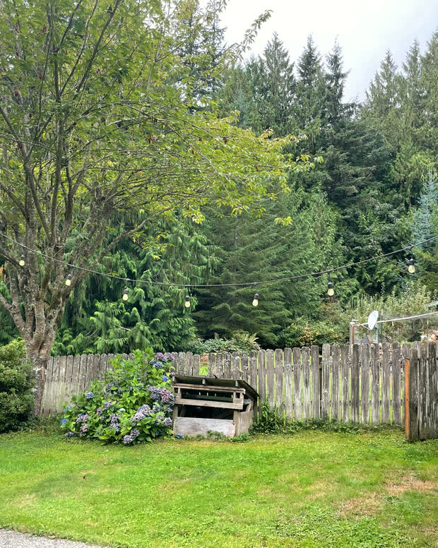 A fenced garden bed with a compost box and a blue planter, below mature deciduous trees.
