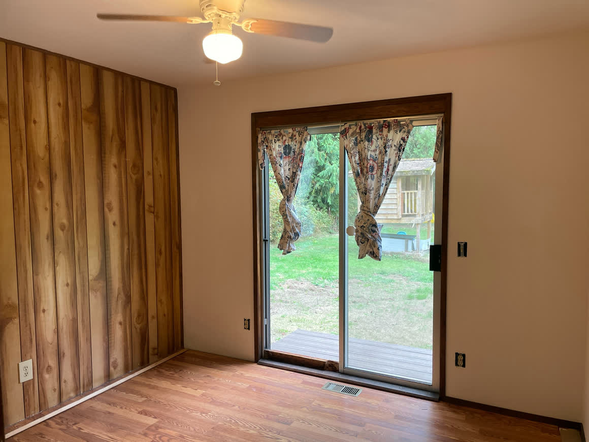 The living room: a knotty-pine panelled wall, hardwood floor, a ceiling fan and a sliding glass door opening onto the deck.