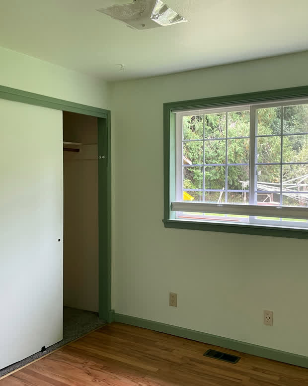 A room with a built-in closet behind white sliding doors, a green-trimmed window and a hardwood floor.