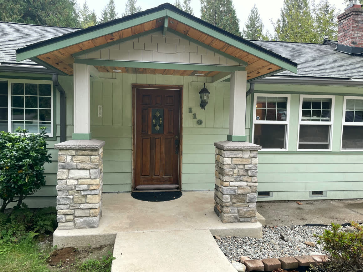 The front entry: a gabled porch carried on stone-veneer columns, a panelled wood door, white-trimmed grid windows and a concrete walkway with river rock alongside.