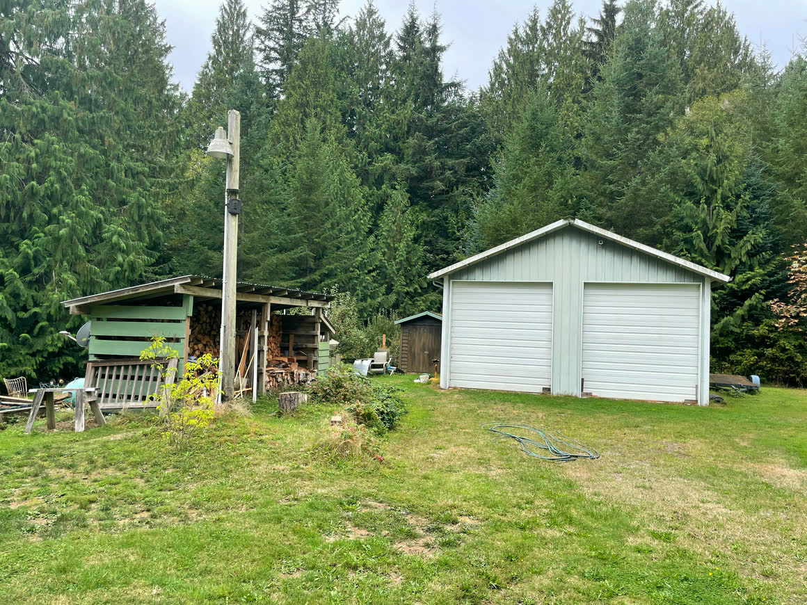 The detached shop: a gable outbuilding with two white overhead doors and vertical siding, with a wood shed and stacked firewood standing alongside it.