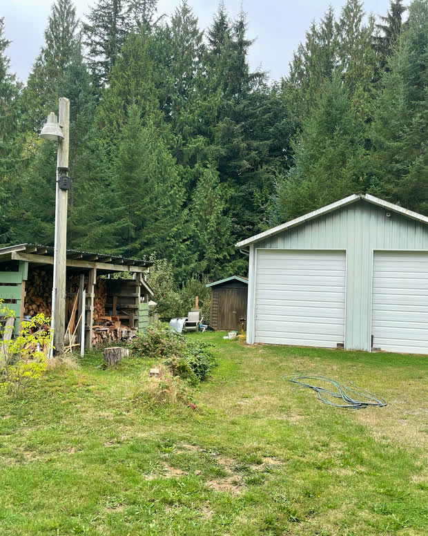 The detached shop: a gable outbuilding with two white overhead doors and vertical siding, with a wood shed and stacked firewood standing alongside it.